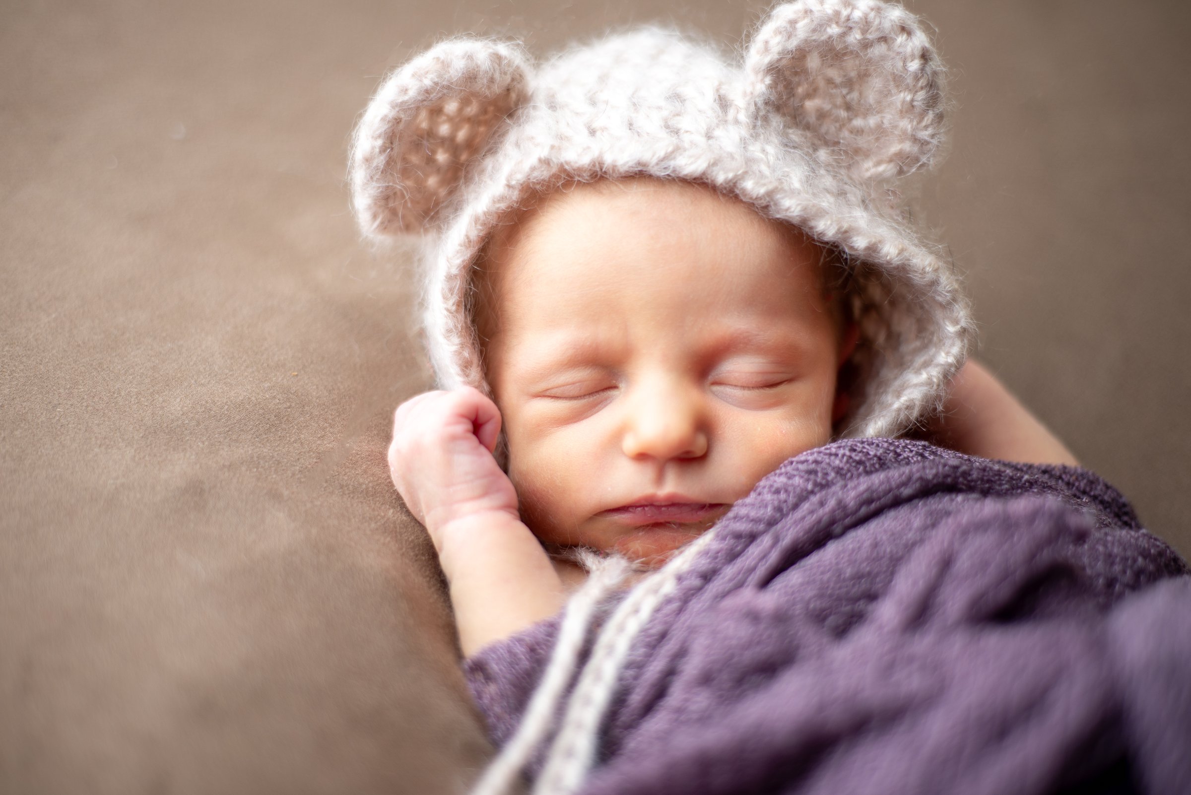 Sleeping newborn wearing a knit bear-ear bonnet, swaddled in a purple wrap